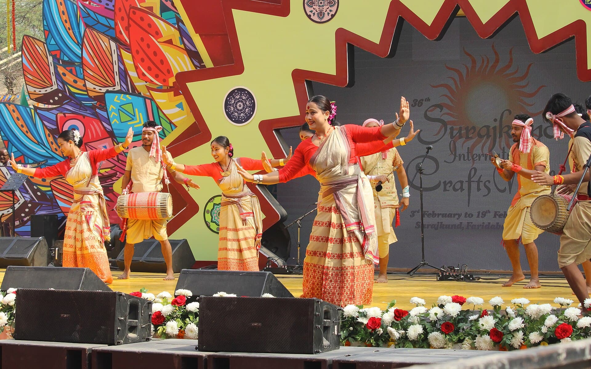 Dancers in red and white mekhela chador and dhuti performing Bihu dance on a stage with dhol players behind