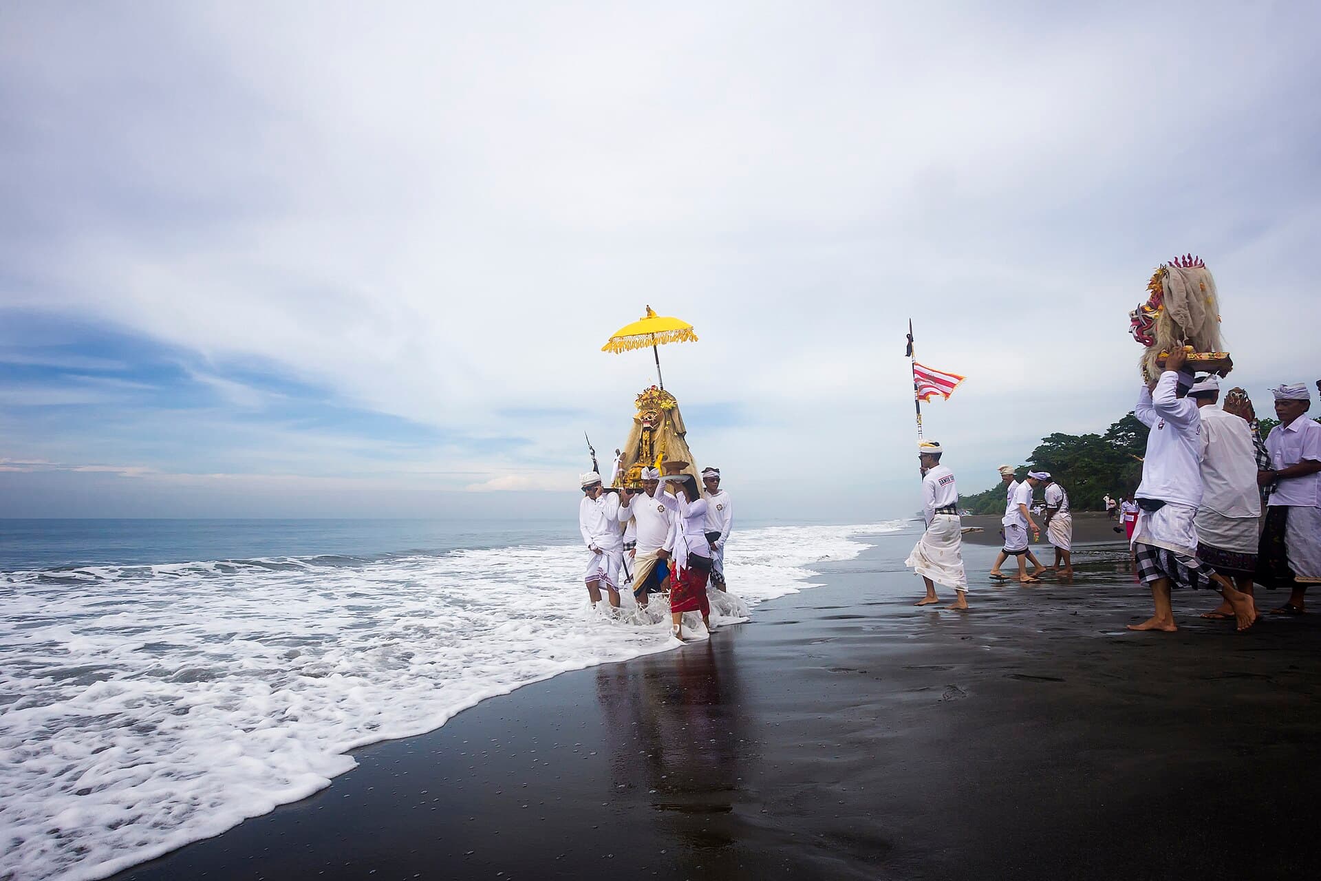 Melasti procession of villagers in white walking onto a Bali beach carrying temple umbrellas and sacred objects