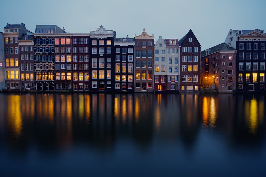 Light artworks reflected in an Amsterdam canal at dusk, with canal houses along the quay