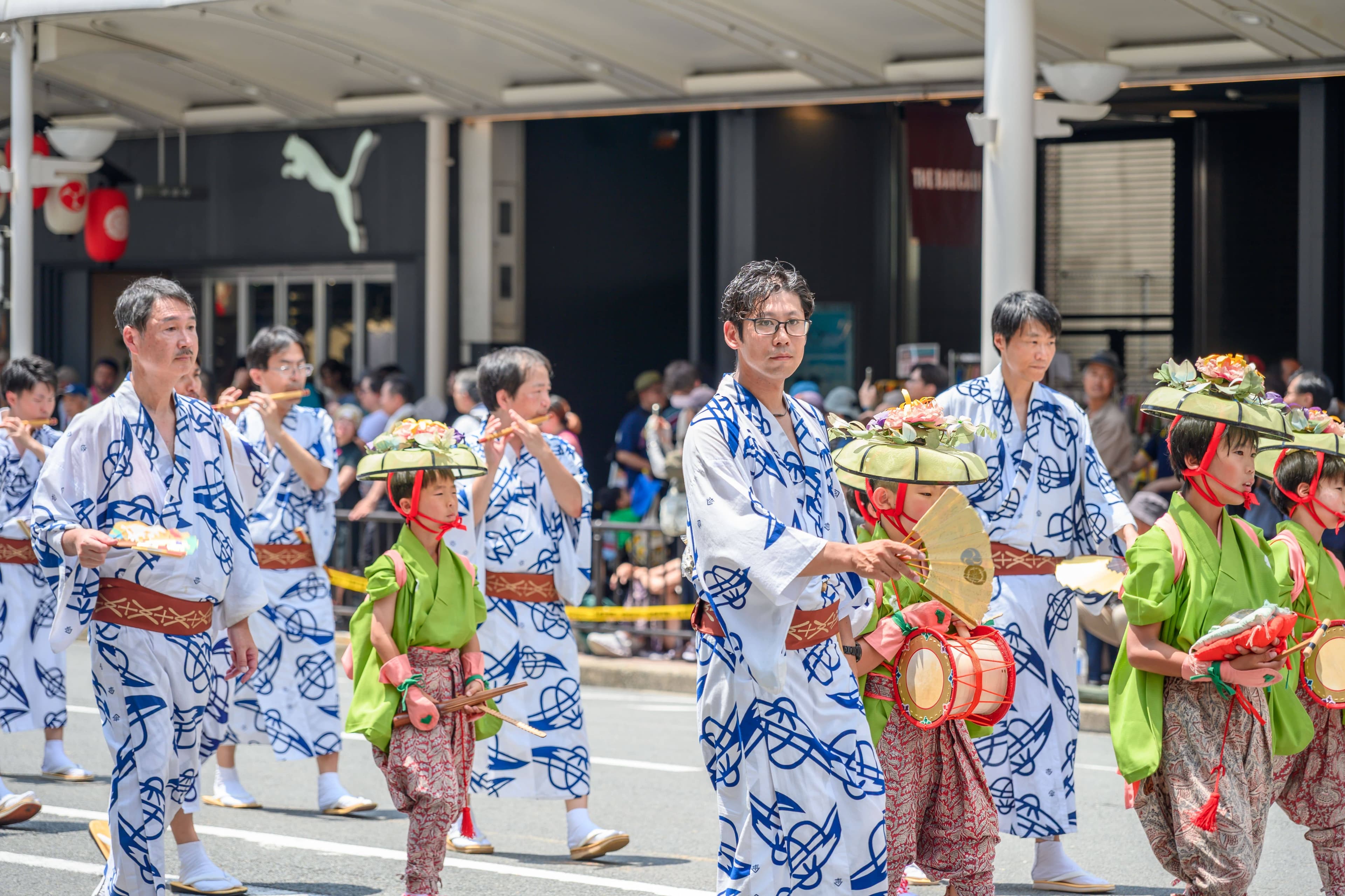 Crowds in yukata holding paper fans walking past lantern-lit yamaboko floats on a hot Yoiyama night in Kyoto